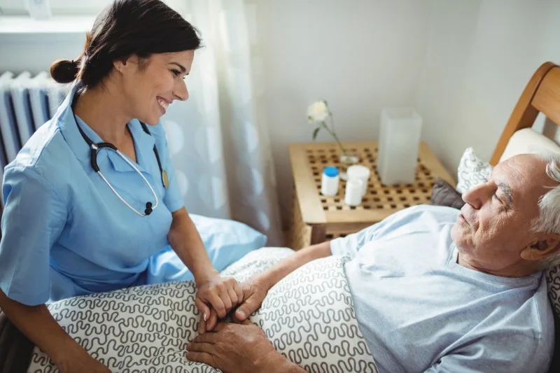Caregiver holding elderly man's hands at bedside