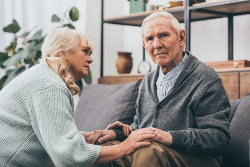 Elderly man seated on sofa with caregiver holding his hands