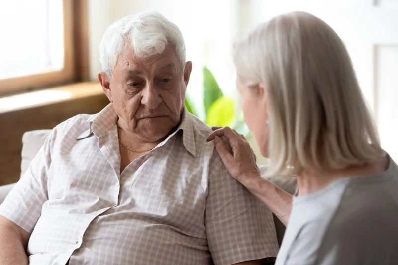Elderly man seated with woman touching his shoulder