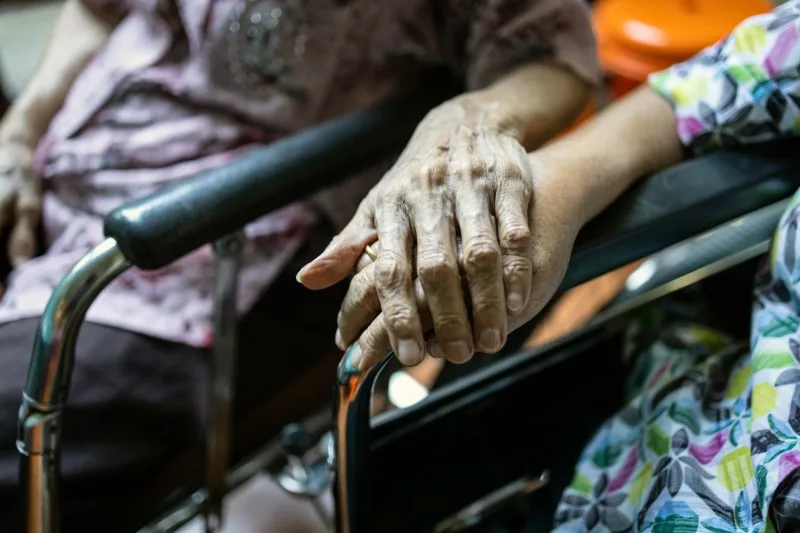 Two elderly hands clasped over a wheelchair arm