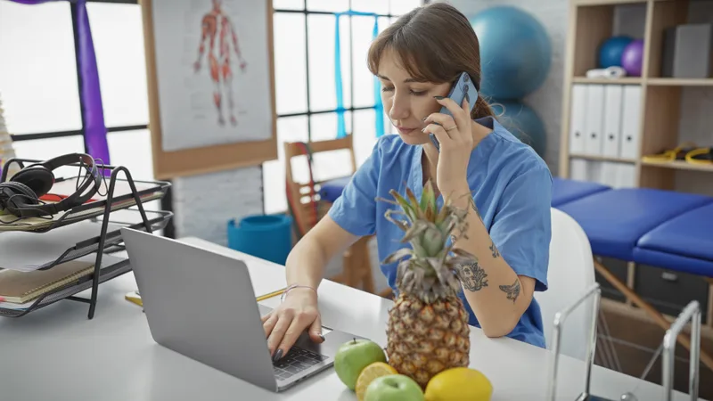 Woman multitasking on a laptop and phone in a busy medical clinic