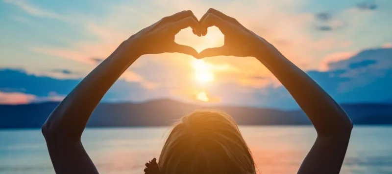Person forming heart shape with hands at sunset