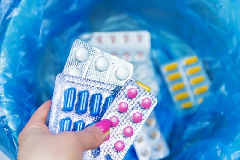 Hand holding blister packs of pills over a blue trash bag